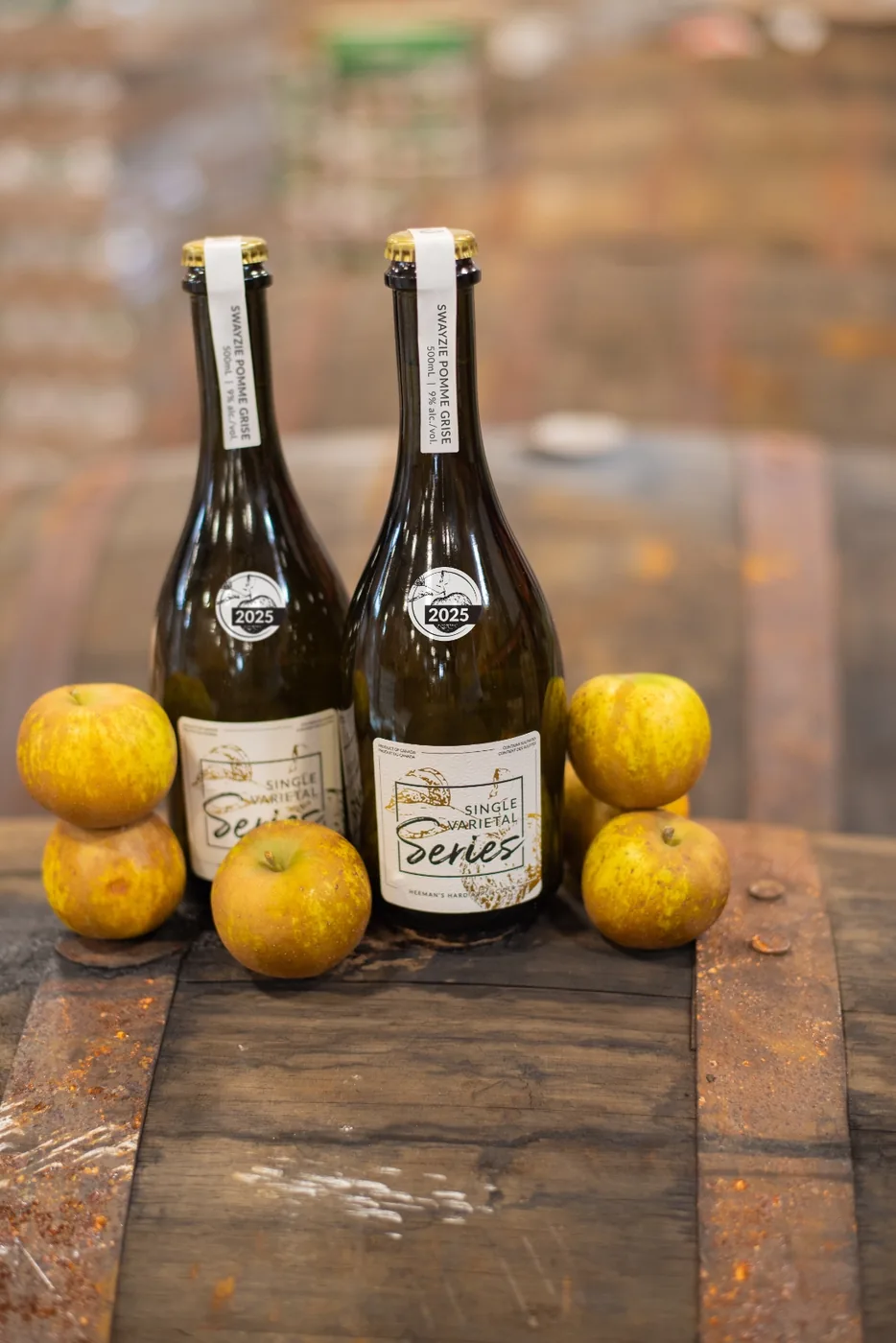 Two bottles of Swayzie Pomme Grise on top of an oak barrel flanked by stacked russeted apples.
