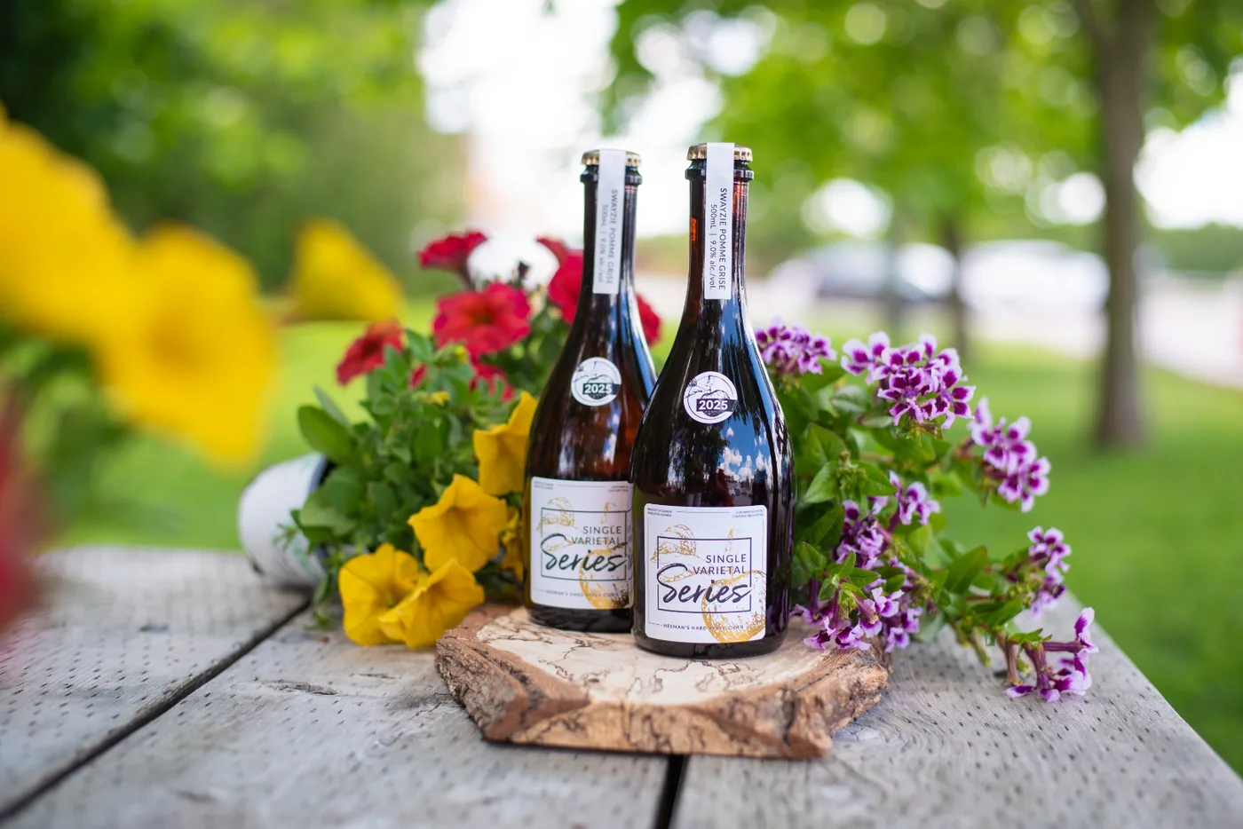 Two bottles of Swayzie Pomme Grise hard cider on a live-edge wood round, surrounded by summer petunias on a weathered picnic table at Heeman's.