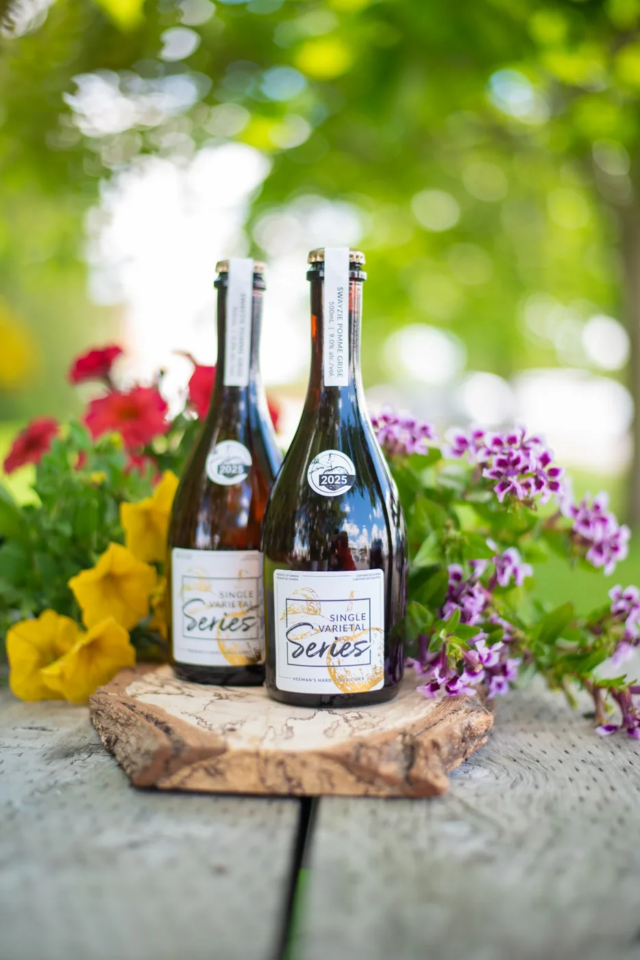 Vertical view of two Swayzie Pomme Grise bottles on a wood round with red, yellow and purple summer blooms behind.