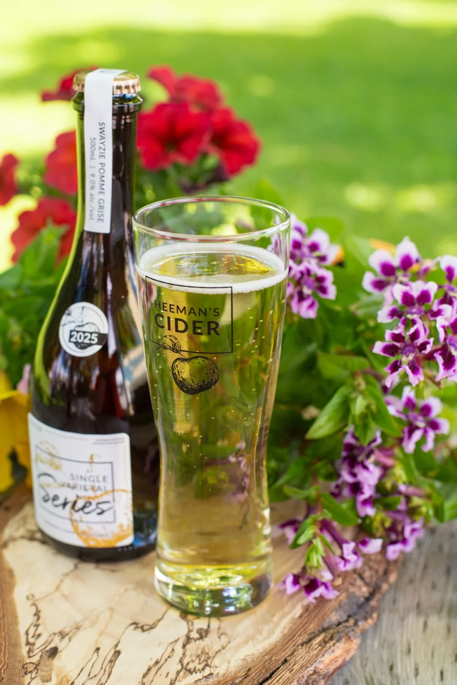 A full branded Heeman's Cider glass of pale golden cider beside a bottle of Swayzie Pomme Grise, surrounded by flowers.