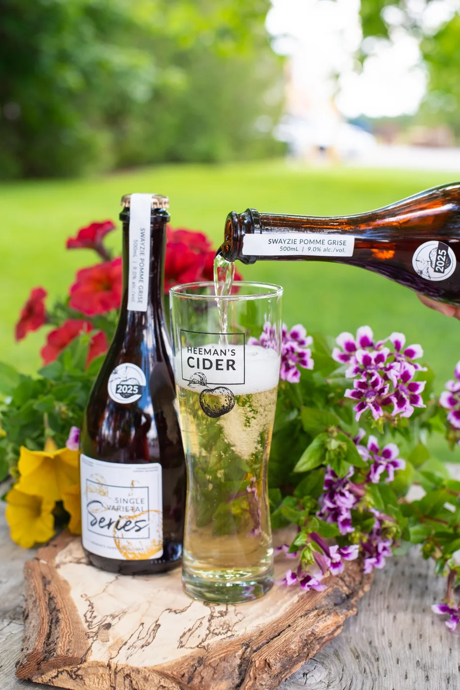 Pale golden cider being poured from a bottle into a branded Heeman's Cider glass on a wood round outdoors.