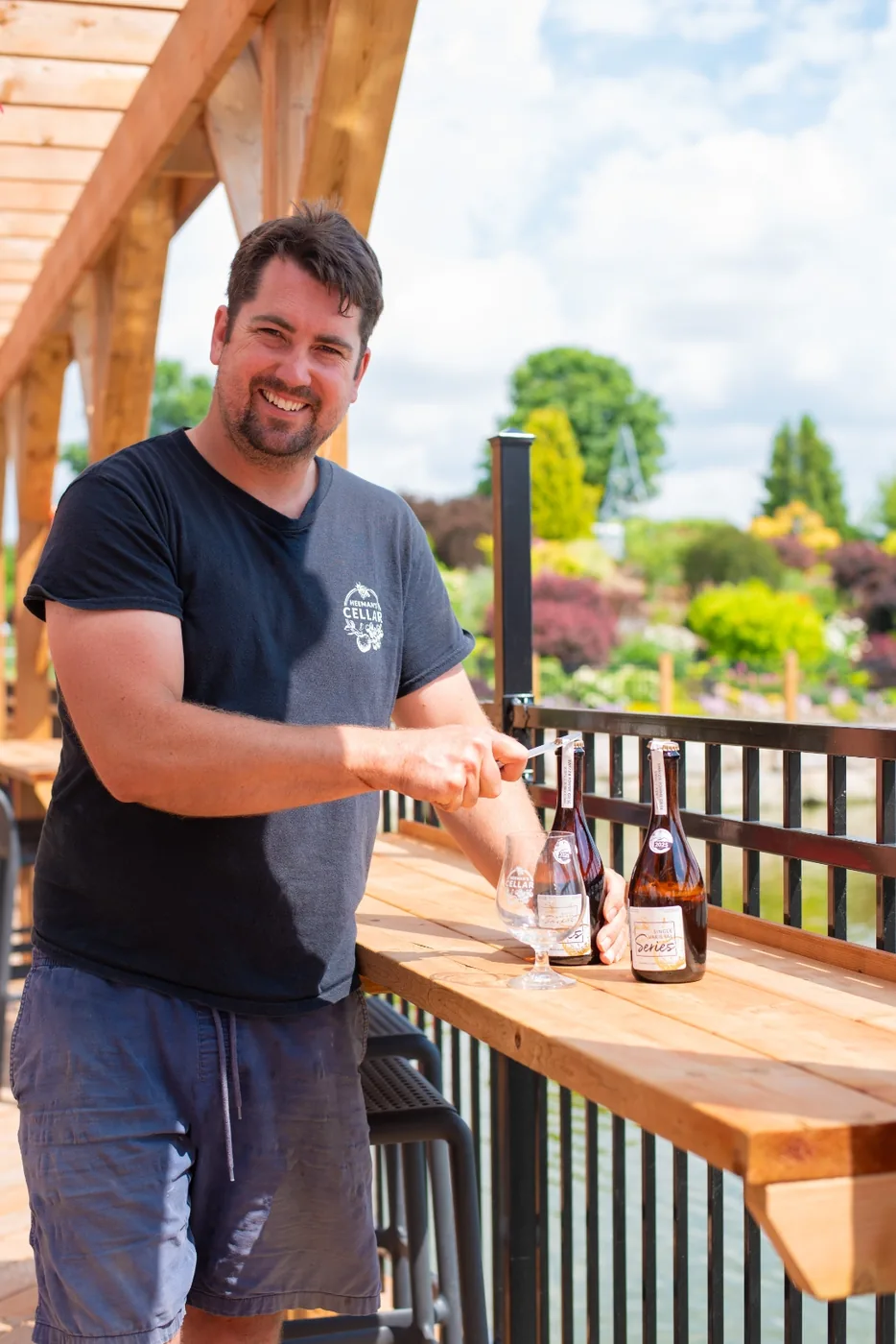 Tom Heeman in a Heeman's Cellar t-shirt opening a bottle at a wooden bar on the Dock, garden beds and pond behind.